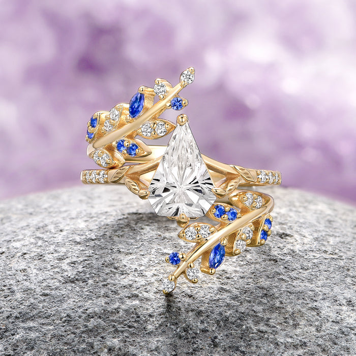 Blue Sapphire & Fantasy Shield Cut Diamond (CZ) Couples Ring Set by Aquamarise, featuring a yellow gold band with a large fantasy shield-shaped clear cubic zirconia center stone, accented with vibrant blue sapphires and sparkling diamond-like CZs in a delicate leaf-inspired design.