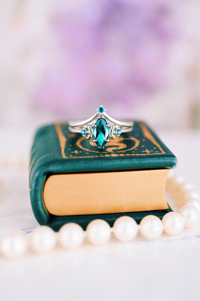 Borealis Forest & Teal Sapphire Couples Ring Set- Northern Lights & Trees featuring a marquise teal sapphire centerpiece with smaller accent stones set in a delicate silver band, displayed on a vintage green book with a pearl necklace, crafted by Aquamarise for Matching Couples' Rings and September Birthstone Jewelry collections.