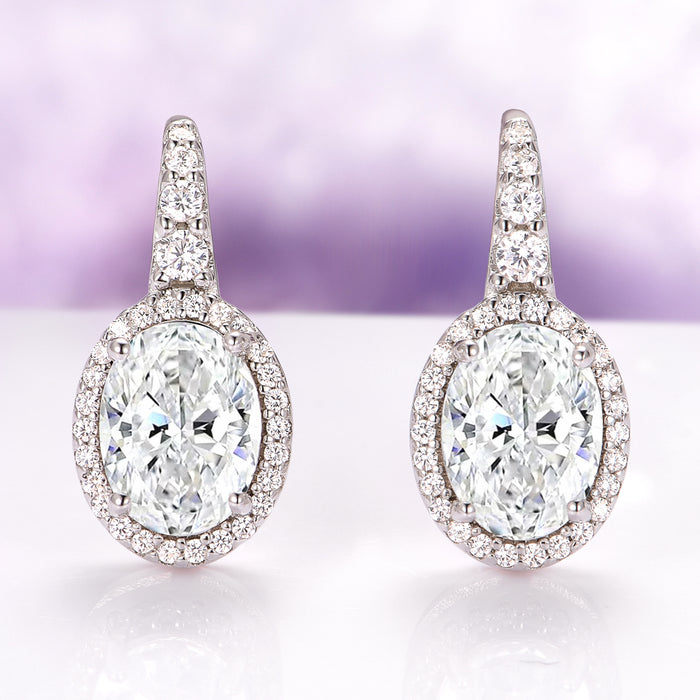 Simulated Diamond Dangle Earrings in Sterling Silver by Aquamarise, featuring oval-cut simulated diamonds surrounded by smaller stones on elegant drop settings against a soft purple background.