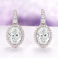 Simulated Diamond Dangle Earrings in Sterling Silver by Aquamarise, featuring oval-cut simulated diamonds surrounded by smaller stones on elegant drop settings against a soft purple background.