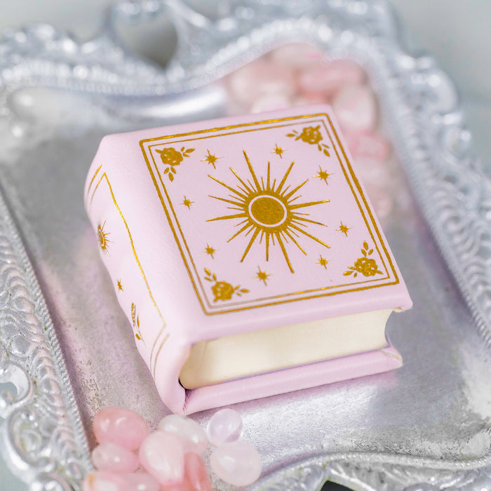 Dawn Court Leather Book Ring Box - Blushing Dawn Edition (Small) by Aquamarise, a soft pink handmade book-shaped ring box with gold celestial design on a silver tray with rose quartz stones.