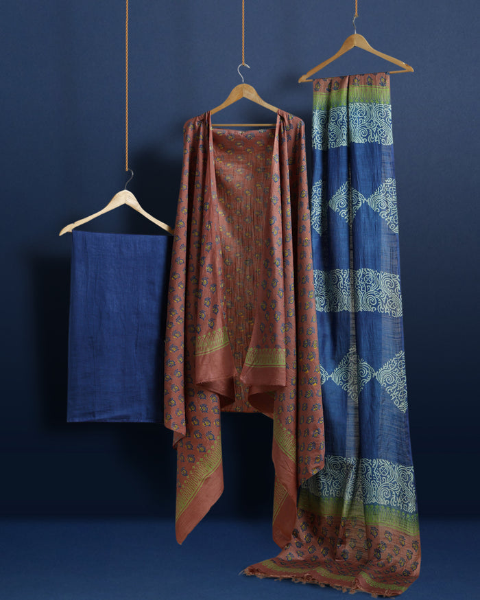 Deep blue katan silk unstitched suit with all-over block-printed floral and diamond motifs, a rust-orange dupatta with wide geometric end borders, and a plain blue bottom.