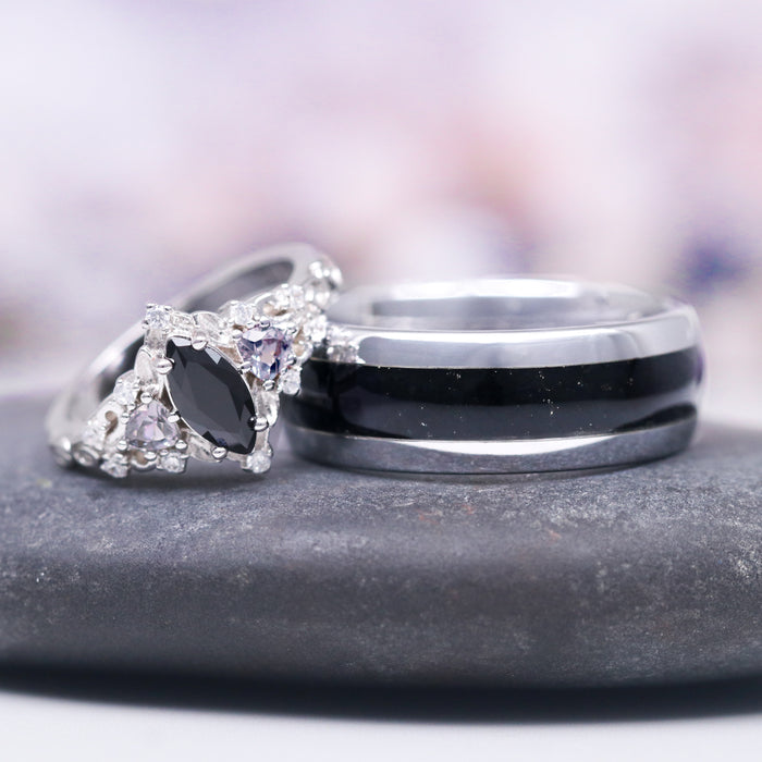 Black Onyx Wedding Band in Tungsten Carbide (8mm) displayed beside an ornate silver ring with black and clear gemstones on a smooth gray stone by Aquamarise.