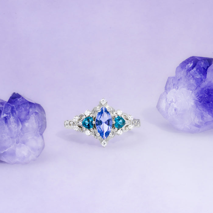 Silver ring with blue gemstone surrounded by purple crystals on a light purple background