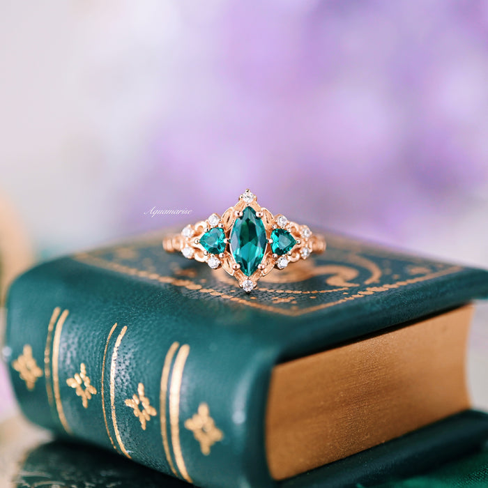 Elain Teal Sapphire Fairy Engagement Ring in 14K Rose Gold Vermeil with marquise and heart-shaped teal sapphires, accented by diamonds, styled on a vintage green book by Aquamarise.