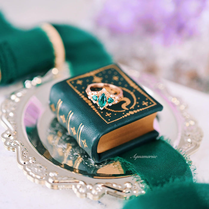 Elain Teal Sapphire Fairy Engagement Ring in 14K Rose Gold Vermeil displayed on a miniature decorative book with green and silver accents by Aquamarise.