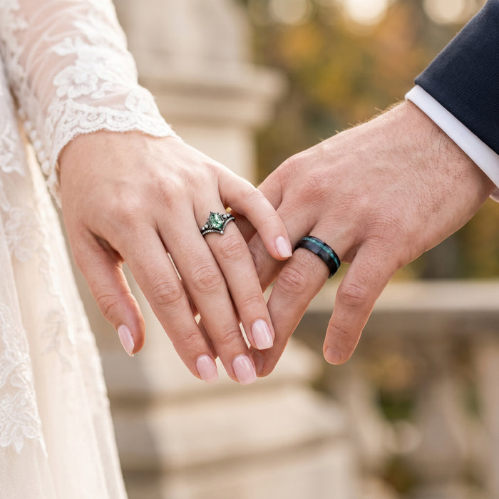Crushed Moss Agate Men's Wedding Band in Black Tungsten (8mm) by Aquamarise, featuring a dark band with green moss agate inlay worn on the groom’s hand beside a bride’s hand with a green gemstone ring.