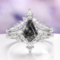 Diamond ring with a central black diamond and pear-shaped side stones on a reflective surface with a blurred purple background.