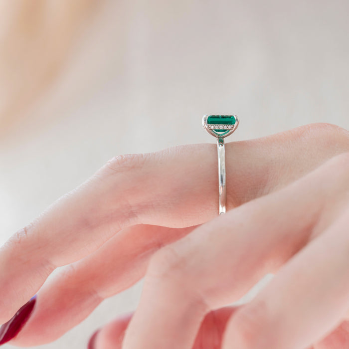 Emerald Engagement Ring in 14K White Gold (Hidden Halo) by Aquamarise Gold, featuring a slender white gold band with a unique emerald-cut lab-grown emerald worn on a delicate finger.