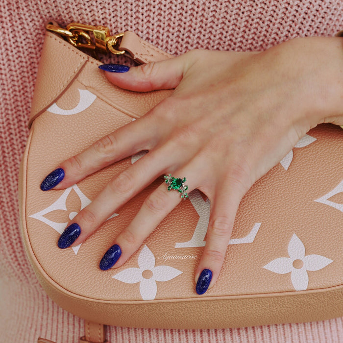 Oval Cut Lab-Grown Emerald Leaf Engagement Ring in 14K White Gold by Aquamarise Gold, featuring vibrant green emeralds and leaf-inspired design on hand with glittery dark blue nails.