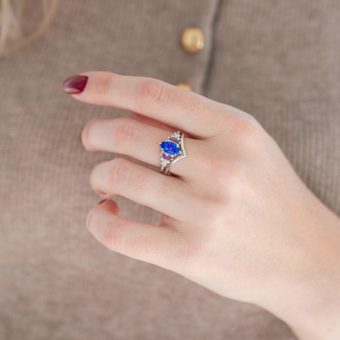 Silver marquise purple and pink opal engagement ring set with marquise pink sapphire and diamond accents paired with a chevron band displayed in hand against a warm brown background