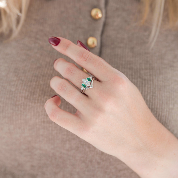 Silver marquise diamond engagement ring set with marquise green emerald and diamond accents paired with a chevron band displayed in hand against a warm brown background