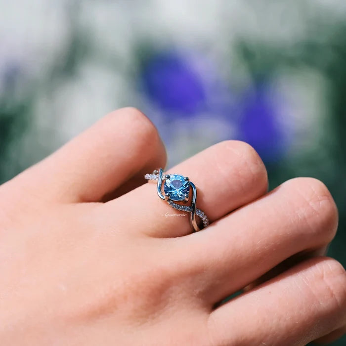 Entwined Aquamarine Ring- Sterling Silver by Aquamarise, featuring a delicate twisting band with pavé accents and a bright blue aquamarine center stone on a hand outdoors.