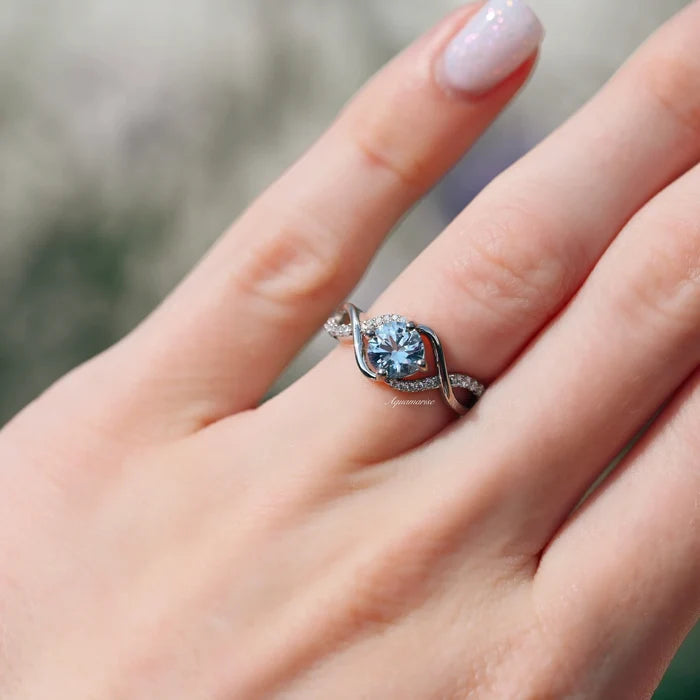 Entwined Aquamarine Ring- Sterling Silver by Aquamarise, showcasing a polished twisting band with pavé accents and a round aquamarine gemstone on a hand outdoors.