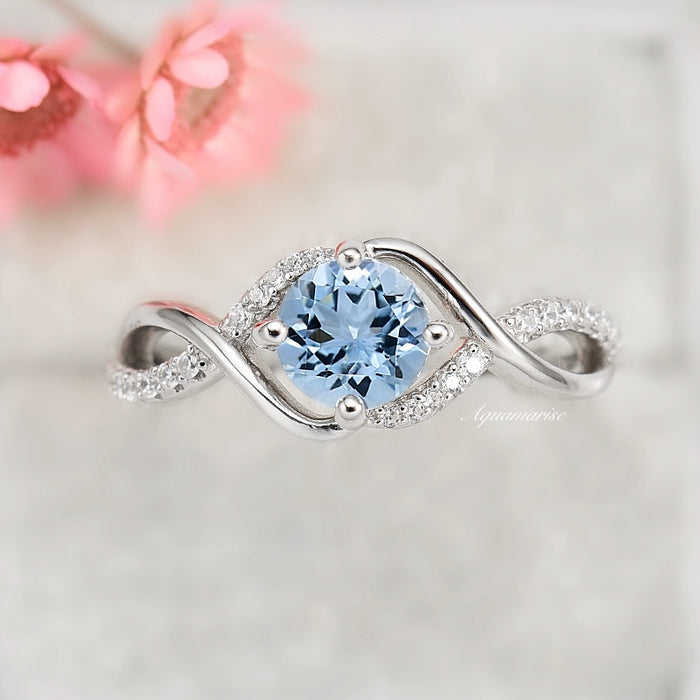 Entwined Aquamarine Ring- Sterling Silver by Aquamarise, featuring a twisting sterling silver band with pavé accents and a round blue aquamarine center stone against a soft blurred background.