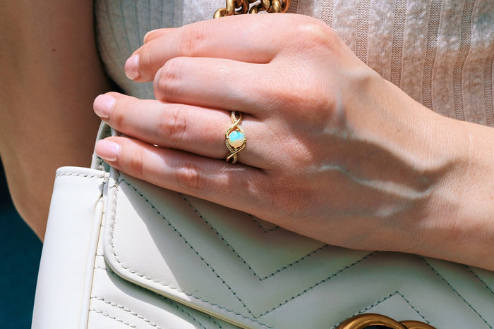 Entwined Natural Australian Opal Engagement Ring in 14K Solid Yellow Gold by Aquamarise Gold, featuring a glowing opal with intertwined band, styled on a hand holding a white quilted purse.