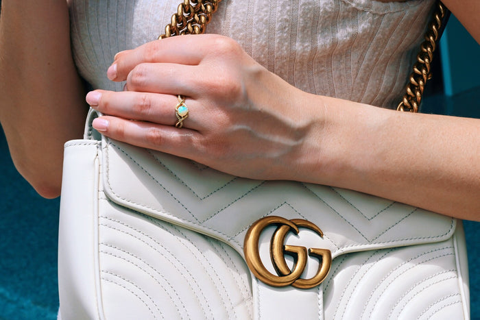 Entwined Natural Australian Opal Engagement Ring in 14K Solid Yellow Gold by Aquamarise Gold, showcasing a glowing opal on a twisted band, worn on a hand holding a white quilted purse.