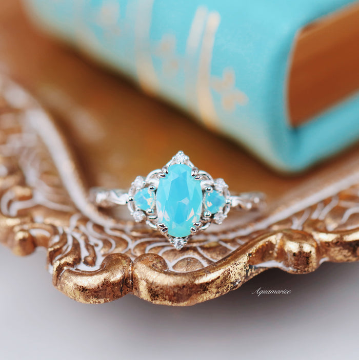 Esme Opalescent Blue Sapphire Mermaid Ring in 925 Sterling Silver by Aquamarise, featuring an oval light blue sapphire with diamond accents on an ornate silver band, styled on a vintage gold tray.