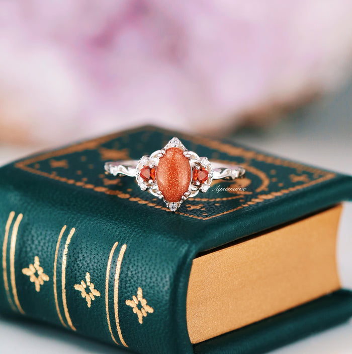 Esme Red Goldstone & Orange Sapphire Ring in Sterling Silver by Aquamarise featuring an oval red goldstone center with sparkling orange sapphires and delicate vintage-inspired silverwork on a green leather book.
