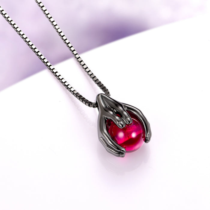 Ethereal Hold Ruby Pendant Necklace in Black Ruthenium by Aquamarise, featuring a black ruthenium-finished sterling silver pendant shaped like two sculpted hands gently cradling a vibrant round ruby, from July Birthstone Jewelry and Lovers of the Dark™ collections.