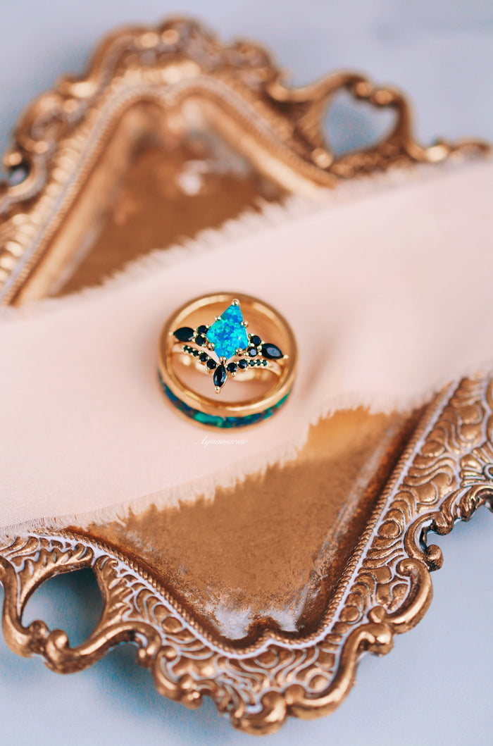 Fire Opal, Meteorite & Starry Night Wedding Band- 8MM Gold Tungsten stacked with a gold ring featuring a vibrant blue-green fire opal center stone and black accent stones, displayed on a soft pink fabric within an ornate gold tray by Aquamarise.