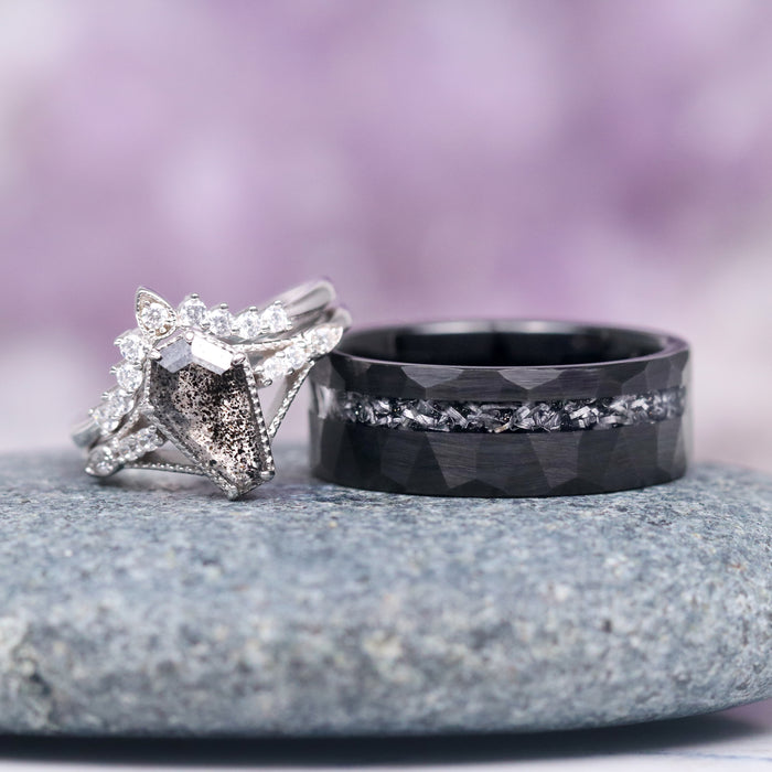 Galaxy Meteorite Hammered Wedding Band- 8MM Black Tungsten by Aquamarise, featuring a faceted black tungsten band with a crushed meteorite inlay, shown beside a white gold ring with a rough diamond center and surrounding white diamonds, styled on a smooth stone surface with soft purple background, part of Men’s Wedding Bands and Meteorite Men’s Wedding Bands collections.