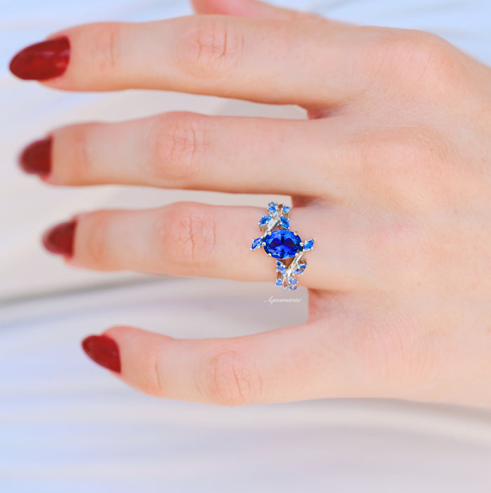 Lab-Grown Oval Cut Cornflower Sapphire Leaf Engagement Ring in 14K White Gold, featuring a deep blue sapphire with delicate leaf design, worn on hand with red nail polish by Aquamarise Gold.