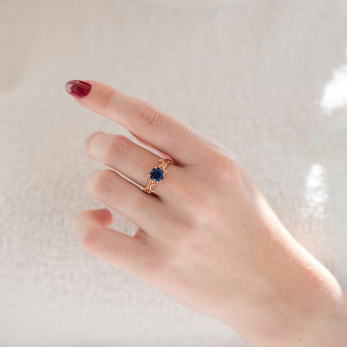Galaxy Sandstone (Starry Night) Leaf Ring- 14K Rose Gold Vermeil by Aquamarise, featuring a sparkling deep blue sandstone in a delicate leaf-inspired rose gold vermeil band on a hand with dark red nail polish.