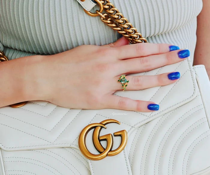 Green Moss Agate & Fire Opal & Gold Leaf Couples Ring Set by Aquamarise on a hand with vibrant blue nails, highlighting the detailed green moss agate and gold leaf design against a white luxury handbag with gold chain straps.
