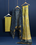 Unstitched Katan silk suit set with deep indigo panels block printed in folk figure silhouettes and geometric shapes, a plain olive green piece, and mustard yellow figurative border bands at the edges and dupatta ends.