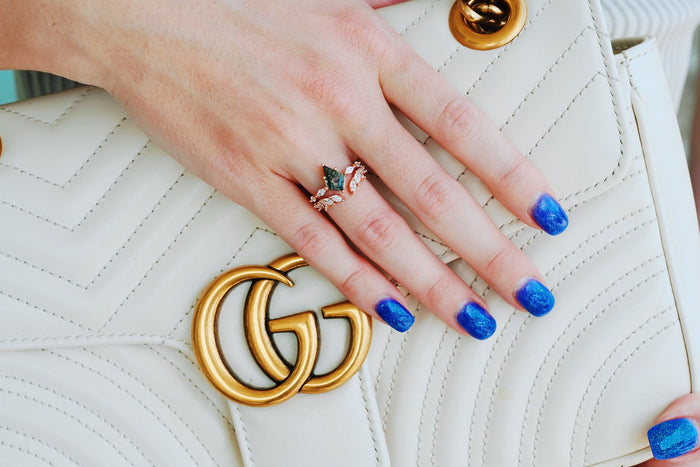Curved Marquise Wedding Band in Solid 14K Rose Gold by Aquamarise Gold, featuring marquise and round stones on hand with blue gradient nails resting on beige quilted bag with gold emblem.