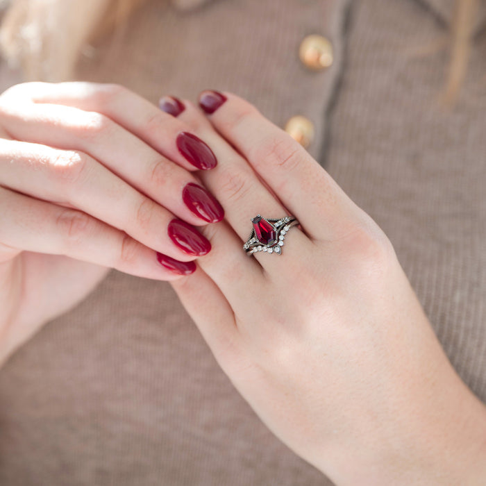 Black coffin-cut dark red garnet engagement ring with diamond accents paired with a wedding band displayed in hand against a warm brown backdrop