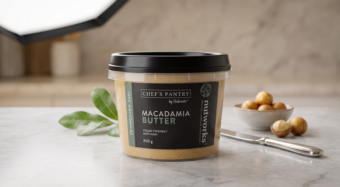 A container  of Nutworks by Chef's Pantry Macadamia Butter sits on a marble countertop with a knife, green leaves, and a small bowl of cashews in a softly lit kitchen setting.