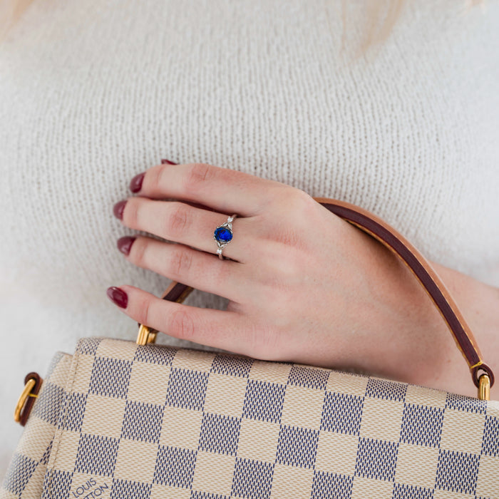 Oval Cut Blue Sapphire Engagement Ring in Sterling Silver by Aquamarise featuring a vibrant blue sapphire with delicate silver detailing, styled on a hand holding a checkered designer handbag.