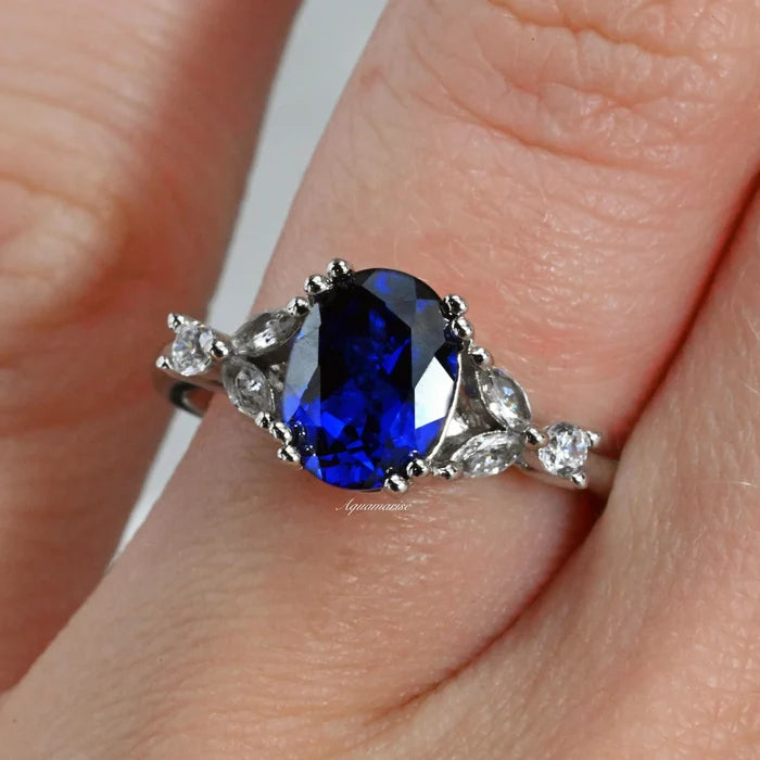 Oval Cut Blue Sapphire Engagement Ring in Sterling Silver by Aquamarise featuring a vibrant oval sapphire center with pear and round diamond accents on a polished silver band worn on a finger.