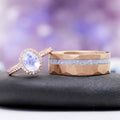Oval Rainbow Moonstone Couples Ring Set by Aquamarise, featuring a rose gold halo ring and a hammered rose gold band with a sparkling moonstone inlay on a black stone with a purple blurred background.