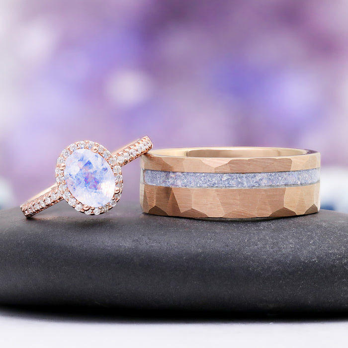 Oval Rainbow Moonstone Couples Ring Set by Aquamarise, featuring a rose gold halo ring and a hammered rose gold band with a sparkling moonstone inlay on a black stone with a purple blurred background.