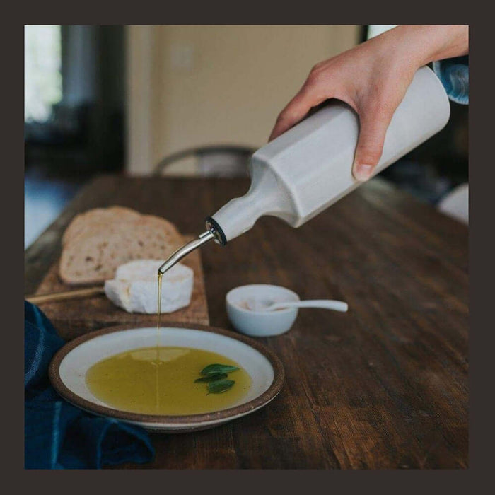 Handcrafted porcelain olive oil dispenser pouring olive oil onto a plate with bread and cheese on a wooden table.