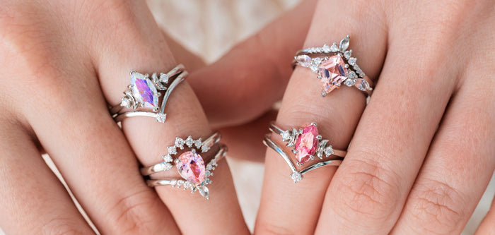 Four silver engagement rings with pink center stones on hands over a light background