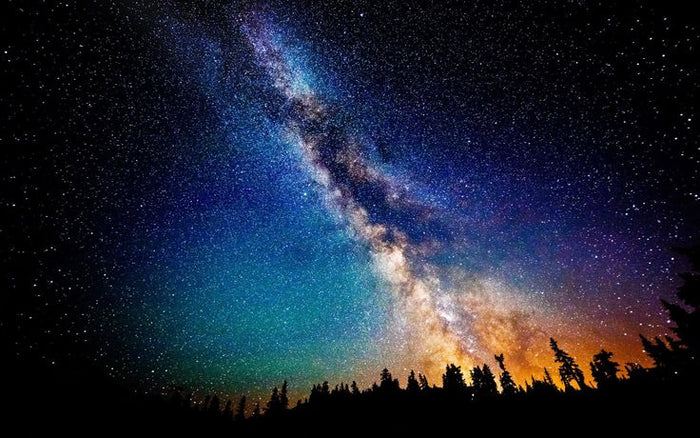 Starry night sky showcasing the vibrant Milky Way galaxy with rich hues of blue, purple, and orange above a silhouette of a forest, evoking a celestial and cosmic atmosphere.