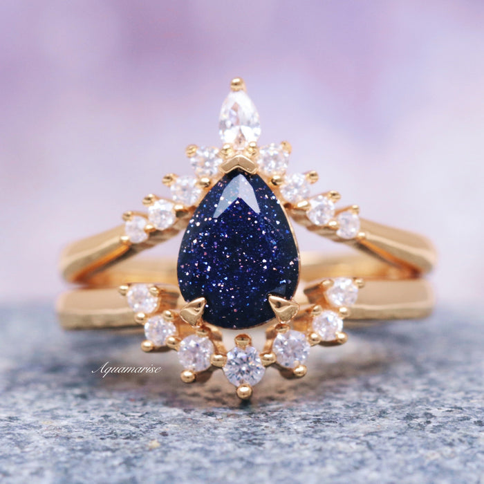A teardrop-shaped blue sandstone gemstone set in a 14K solid yellow gold ring, with smaller clear stones around it, presented on a gray surface.