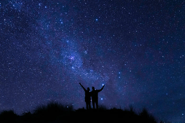 Starry night sky filled with countless stars and the Milky Way galaxy visible above two silhouetted figures on a hill holding a glowing light, evoking a sense of wonder and celestial beauty.