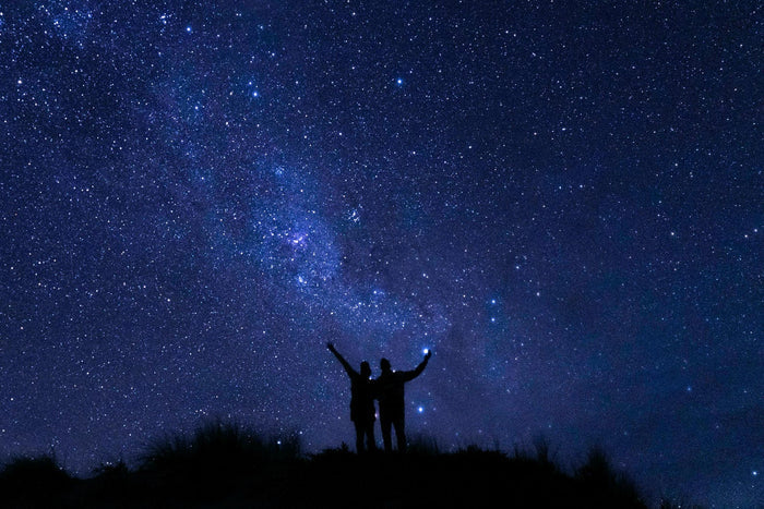 Silhouetted couple standing on a hill under a vast starry night sky filled with countless bright stars and a visible Milky Way, evoking the celestial inspiration behind the Starry Night & Trees Couples Ring Set by Aquamarise.