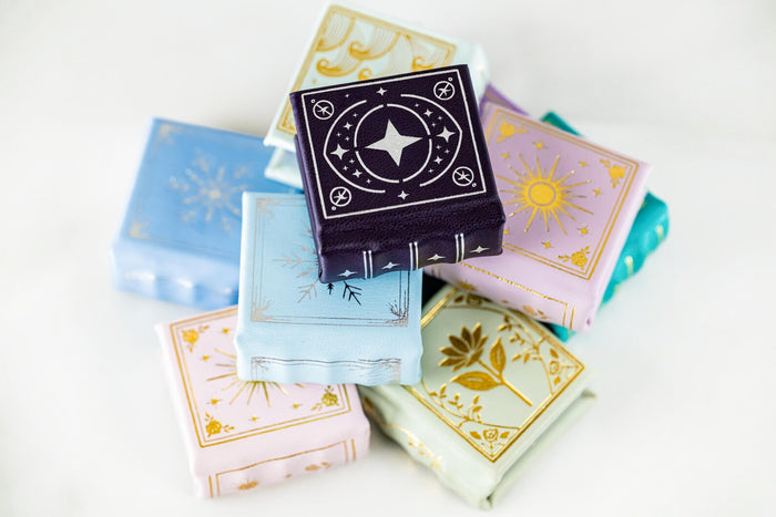 Pile of colorful miniature leather books with gold embossed fantasy designs, featuring the Summer Court Handmade Leather Book Ring Box- Seaglass Glow Edition (Small) by Aquamarise.