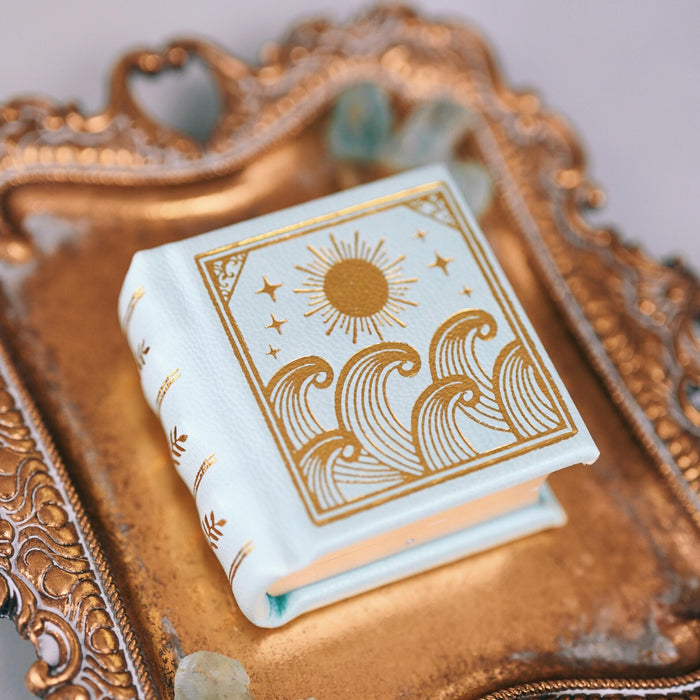 Summer Court Handmade Leather Book Ring Box- Seaglass Glow Edition (Small) resting on ornate gold tray, featuring pale sea-green leather and gold sun and wave design by Aquamarise.