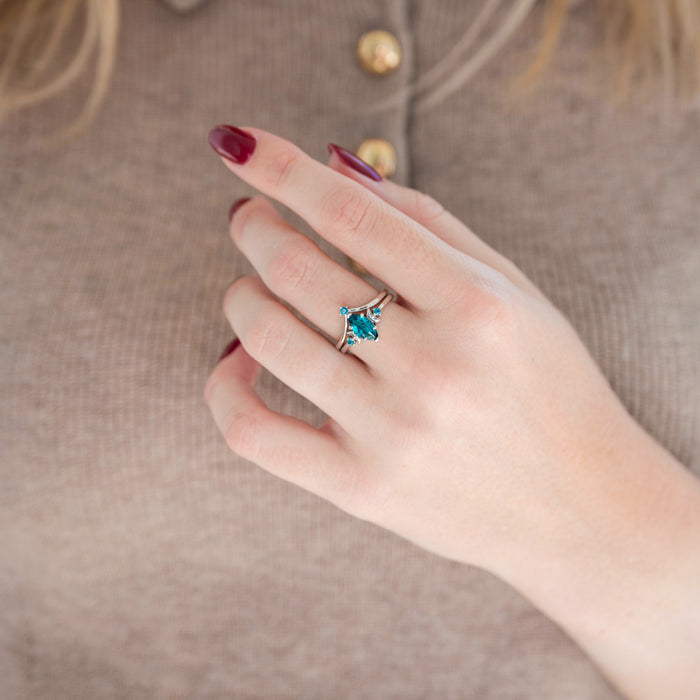 Silver marquise cut teal sapphire engagement ring with crescent moon motif and chevron wedding band held in hand against warm brown background