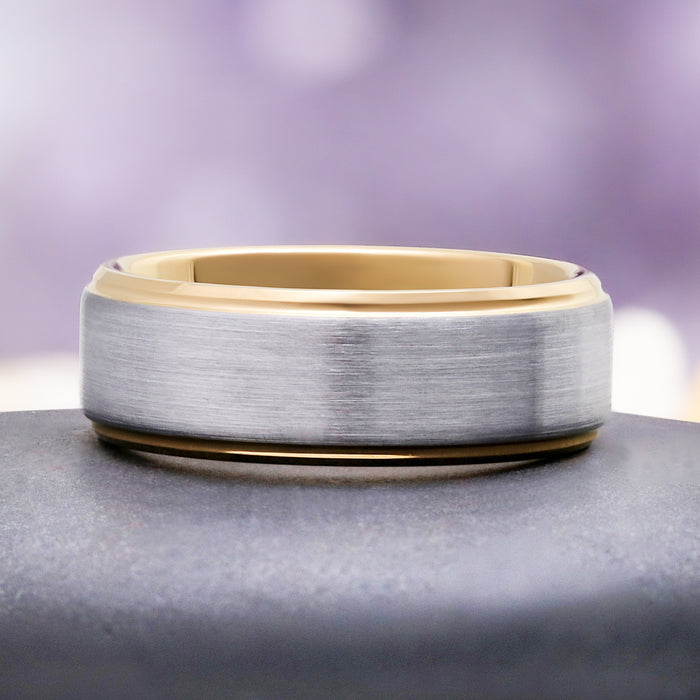 Two-Tone Silver & Gold Men's Wedding Band in Tungsten (8mm) by Aquamarise, a brushed silver band with polished gold stepped edges on a dark stone pedestal.