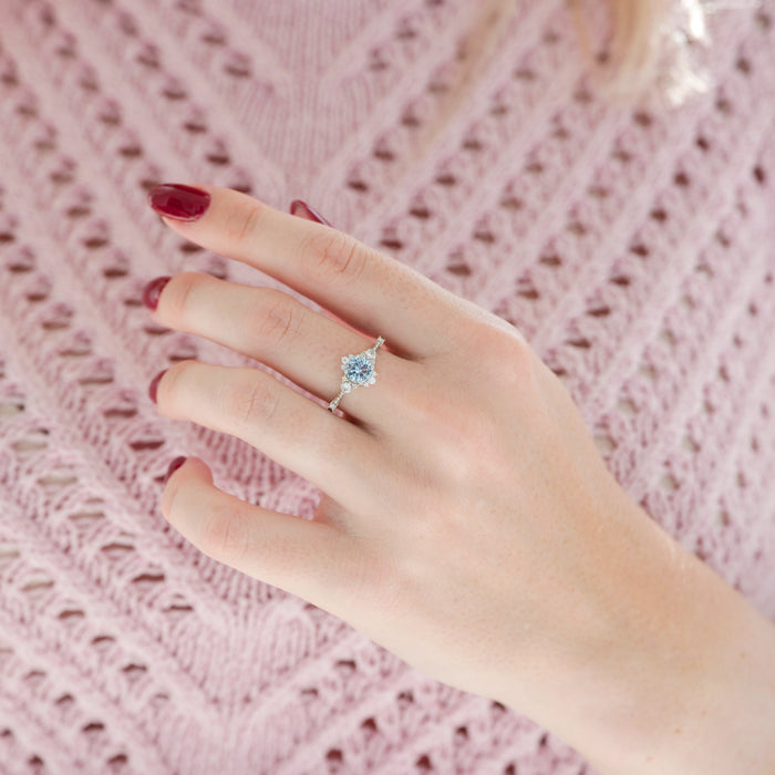 Vintage Round Cut Aquamarine Ring in Sterling Silver by Aquamarise featuring a dainty round aquamarine center set on a delicate band, styled on a hand with burgundy nails and a pink knit sweater.