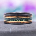 An 8MM two-tone gold and black tungsten wedding band with an inlay of whiskey barrel wood and moss agate stone, displayed against a gradient purple background.