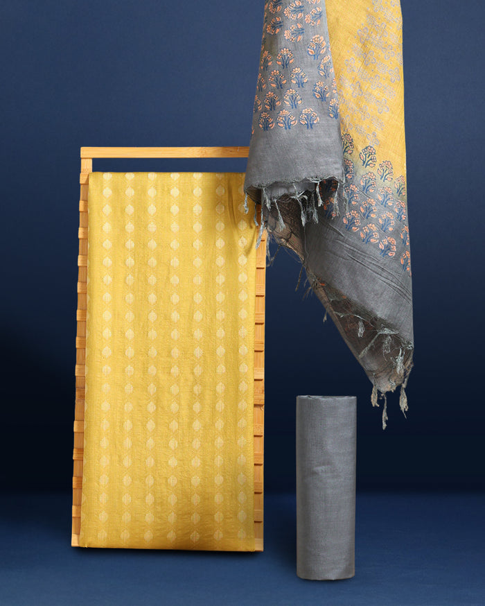 Unstitched yellow Katan silk three piece with small grey ovoid block printed motifs, a densely printed bottom, a sparsely printed top and a dupatta with a floral bordered panel and frayed fringe ends.